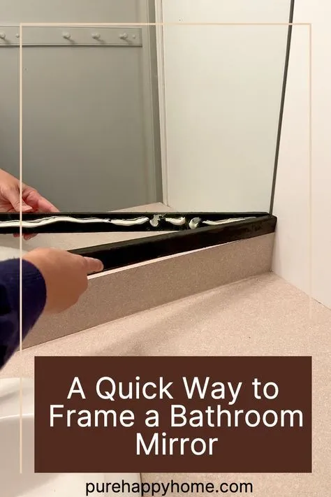 Hands applying a wavy bead of white adhesive to the back of a black mirror frame piece, positioned at a bathroom vanity…